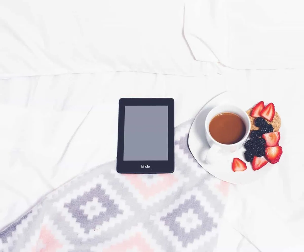 Een Kindle ligt uit op een bed met ernaast een bordje met aardbeien, bosbessen en een kop koffie
