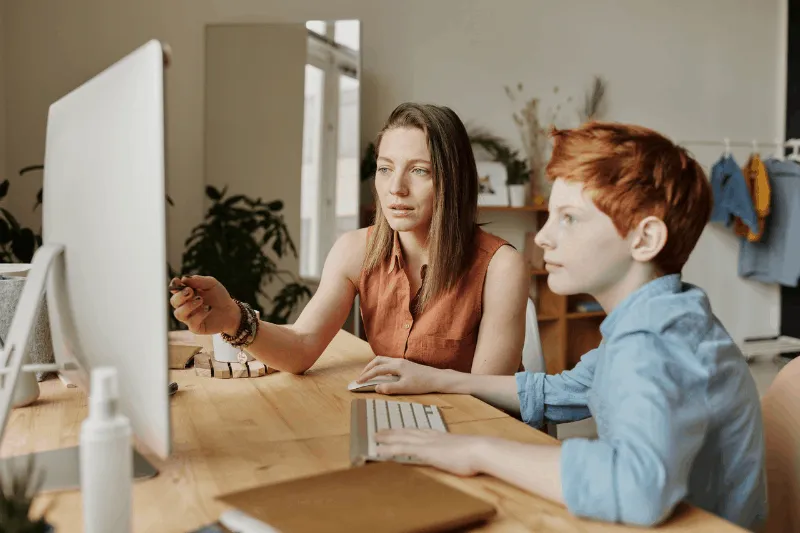 Moeder en zoon zitten achter de computer waar moeder naar iets op het scherm wijst