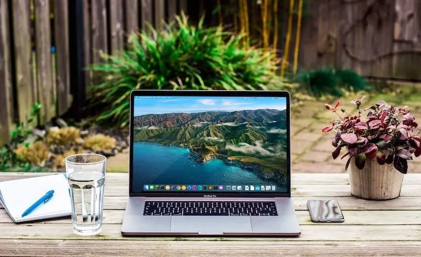 Opengeklapte laptop met middelgroot schermformaat. Staat op een tafel in de tuin met een glas, notitieblok met pen, smartphone en bloemetje ernaast.